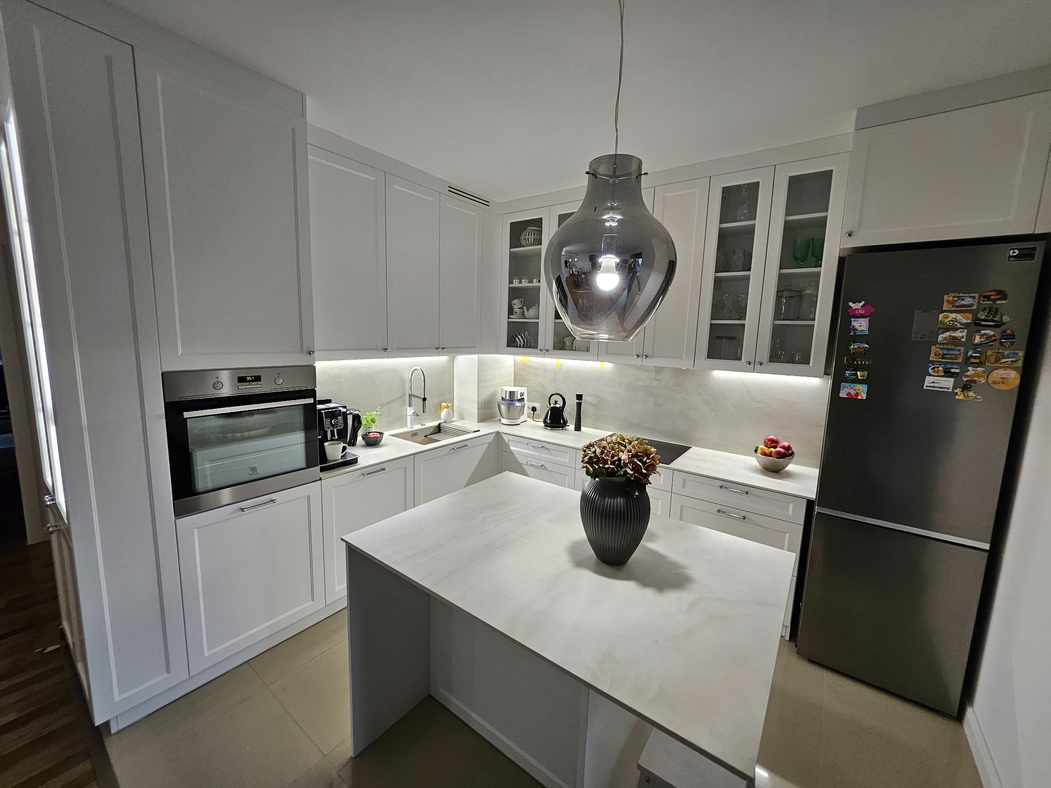 Nowoczesna, jasna kuchnia z białymi szafkami, wyspą z blatem imitującym marmur i designerską lampą wiszącą. Na wyspie wazon z hortensjami. W tle widoczny sprzęt AGD i akcesoria kuchenne.