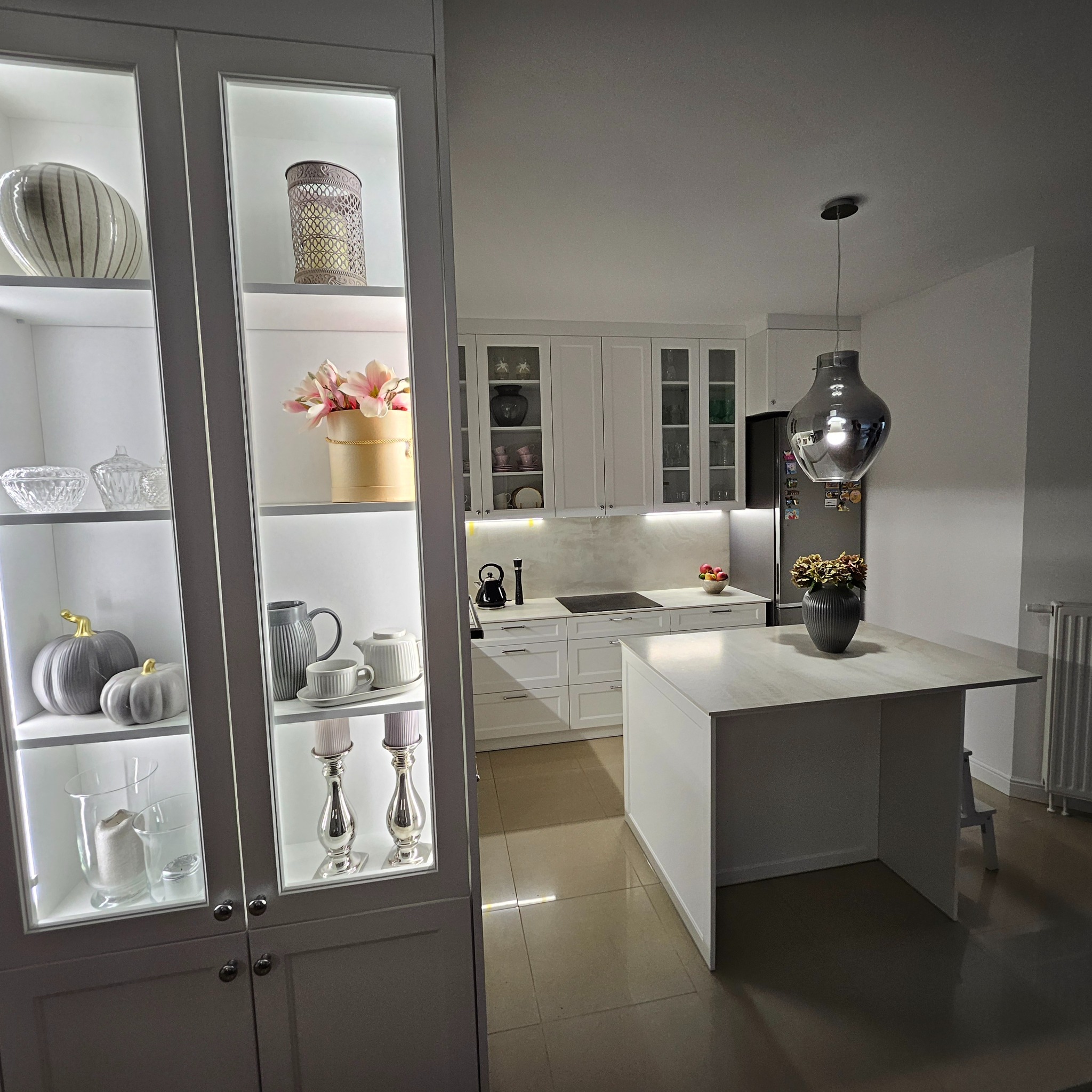 Nowoczesna biała kuchnia z wyspą, oświetlonymi witrynami z dekoracjami i designerską lampą wiszącą.
