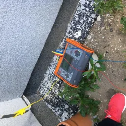 Urządzenie pomiarowe MFT 530-IT z przewodami (czerwonym, niebieskim, żółtym) podłączonymi do ściany budynku, obok widoczna czerwona sportowa podeszwa buta.