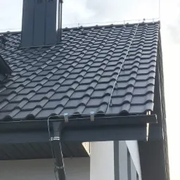 Szary dach domu z ceramiczną dachówką, komin wentylacyjny i system ochrony odgromowej z widocznymi uchwytami mocującymi przewody.