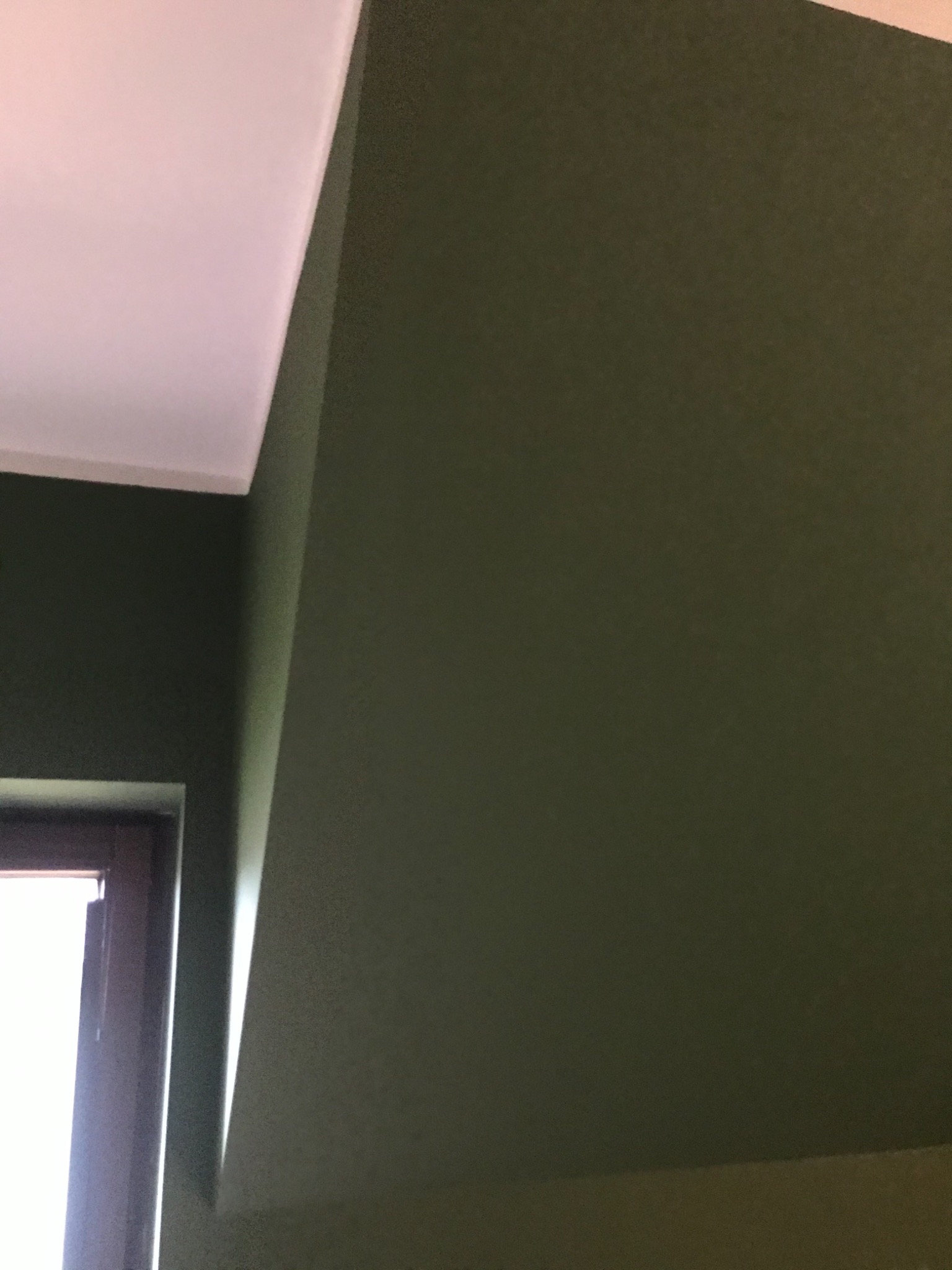Fragment wnętrza z oknem i ścianą pomalowaną na oliwkowy kolor, widoczny fragment sufitu.