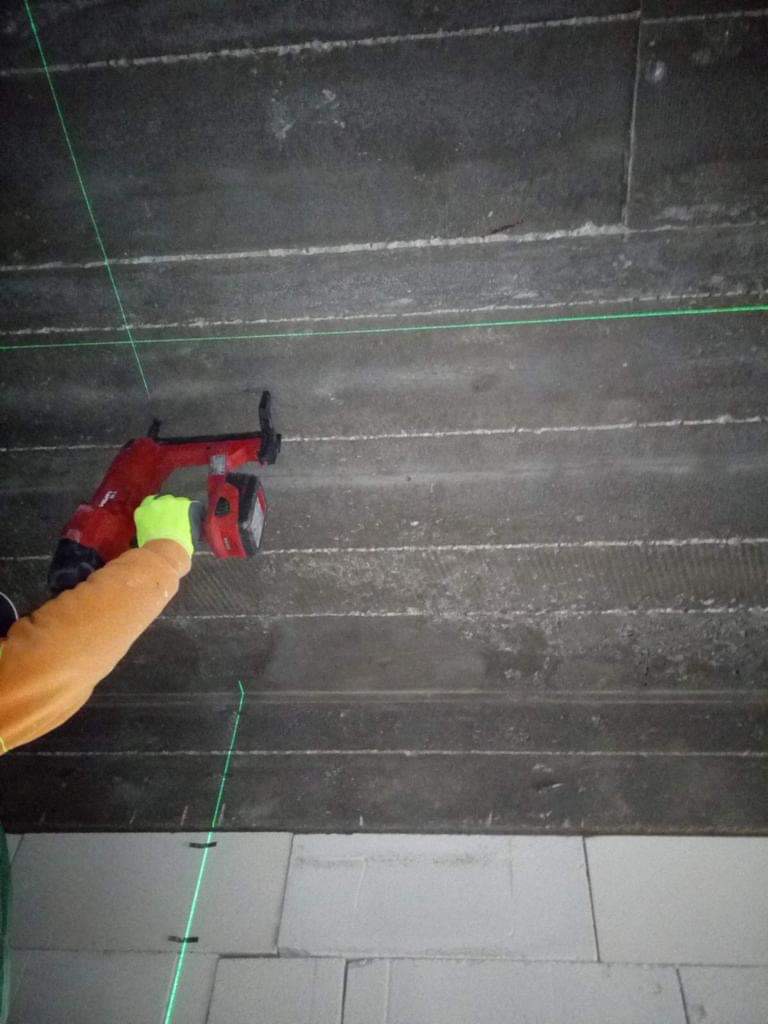 Ustawianie poziomów laserem krzyżowym na betonowym suficie podczas montażu instalacji elektrycznej, widoczna ręka w roboczej rękawicy i czerwony laser.