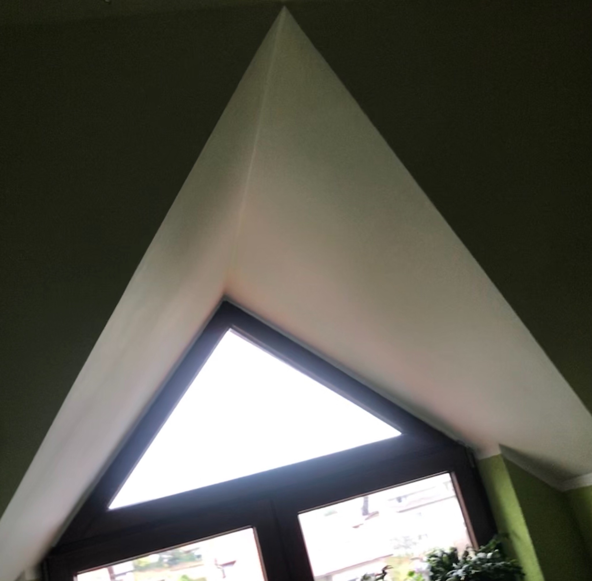 Trójkątne okno dachowe z drewnianą ramą widziane od wewnątrz, z widokiem na zewnątrz i zielonymi ścianami po bokach.