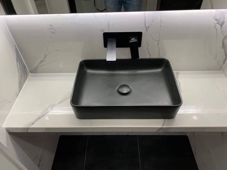 Nowoczesna, prostokątna, czarna umywalka nablatowa z chromowaną baterią na blacie z białego marmuru z szarymi żyłami, podświetlona listwą LED, czarne płytki podłogowe.