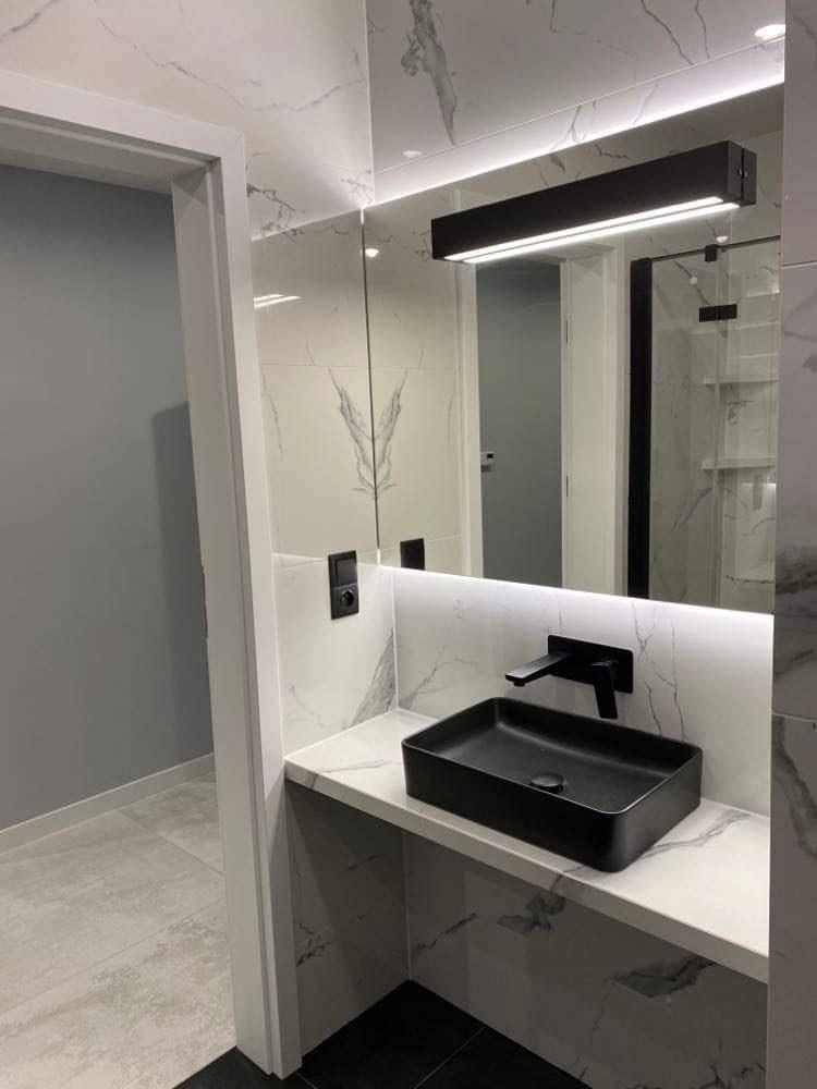 Nowoczesna łazienka z czarną, prostokątną umywalką nablatową i baterią, białe marmurowe płytki na ścianach i blacie, podświetlane lustro z czarną lampą, widoczny fragment kabiny prysznicowej...