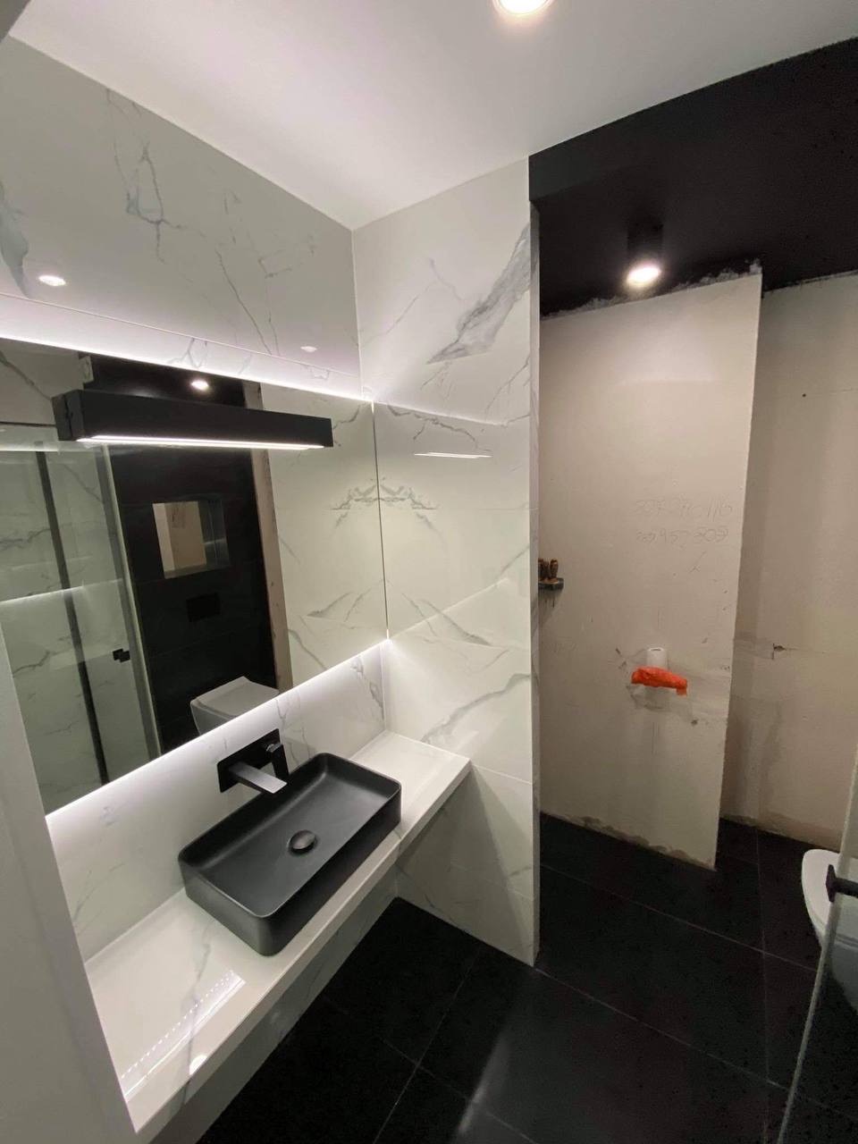 Częściowo wykończona łazienka z czarną, prostokątną umywalką nablatową i baterią, białe płytki imitujące marmur na ścianach, czarna podłoga z płytek. Widoczny fragment ściany przygotowanej...