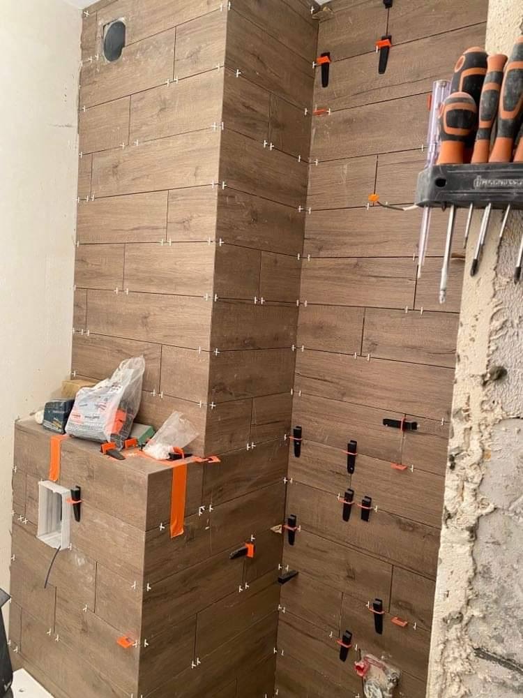 Montaż płytek imitujących drewno na ścianie i obudowie kominka, z użyciem klipsów poziomujących i krzyżyków dystansowych. Widoczne narzędzia i materiały budowlane, surowa ściana w tle.