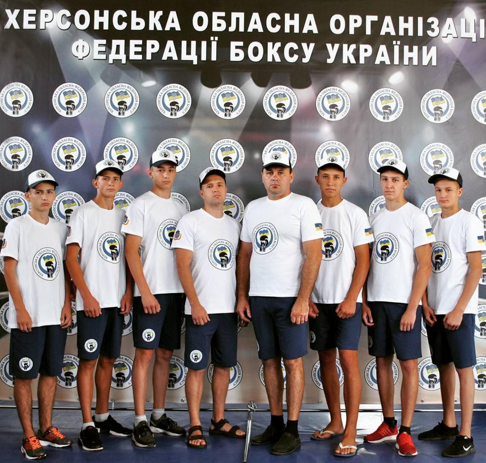 Grupa młodych bokserów w białych koszulkach z logo federacji i granatowych spodenkach, stojących na tle baneru z logo federacji bokserskiej Ukrainy.