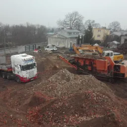 Rozbiórka budynku w toku: ciężarówka Scania z naczepą, koparka VTACHI i kruszarka gruzu na placu budowy, w tle budynki i stos gruzu.