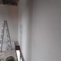 Świeżo nałożone, gładkie tynki na ścianie w surowym wnętrzu, widoczna drabina malarska i wiadro z resztkami zaprawy.