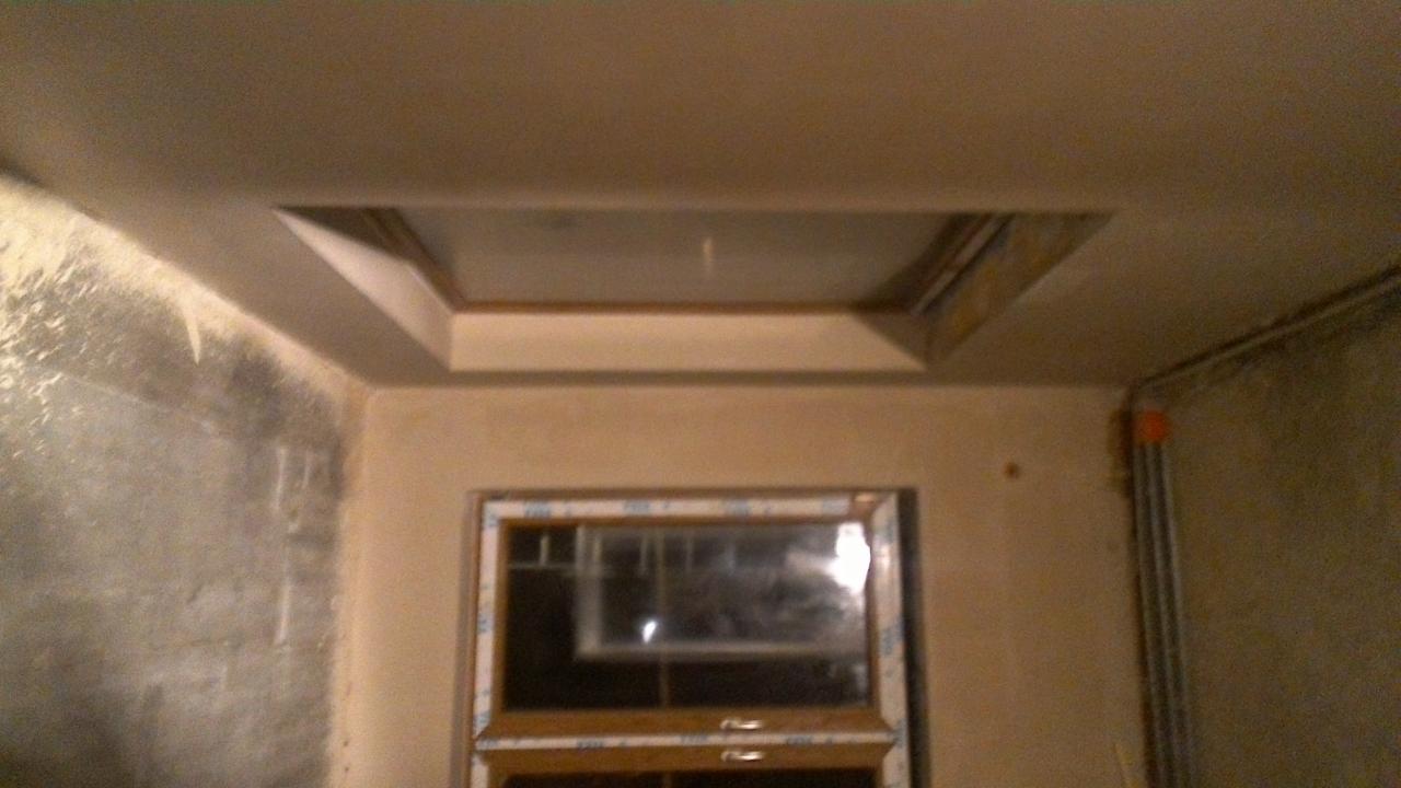 Wnętrze w trakcie prac wykończeniowych: zamontowane okno, ściany częściowo pokryte tynkiem, sufit z dekoracyjnym obniżeniem.