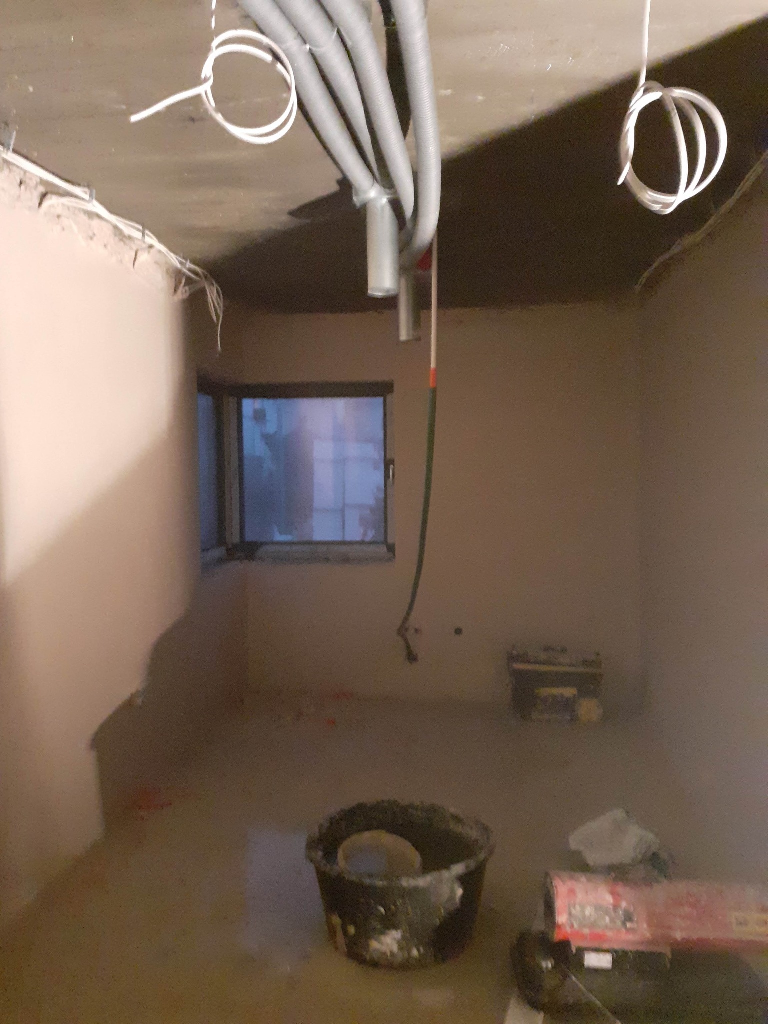 Wnętrze w trakcie prac wykończeniowych: ściany po nałożeniu tynku, widoczne przewody elektryczne pod sufitem, okno, wiadro i maszyna do tynkowania.