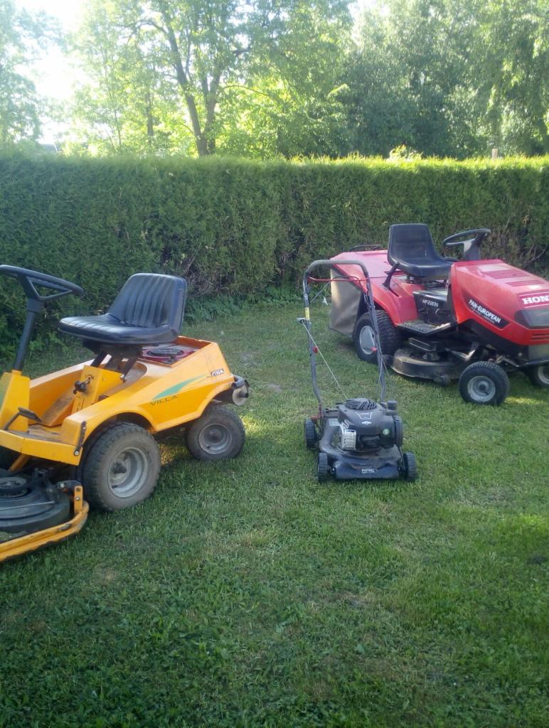 Żółta kosiarka traktorowa Villa, czarna kosiarka spalinowa i czerwona kosiarka traktorowa Honda na trawniku przed żywopłotem.