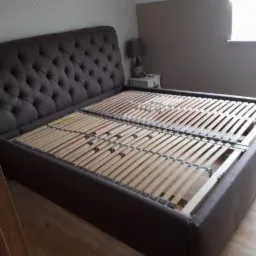 Brązowe łóżko z tapicerowanym zagłówkiem w stylu Chesterfield i drewnianym stelażem, widoczne w pokoju z oknem.