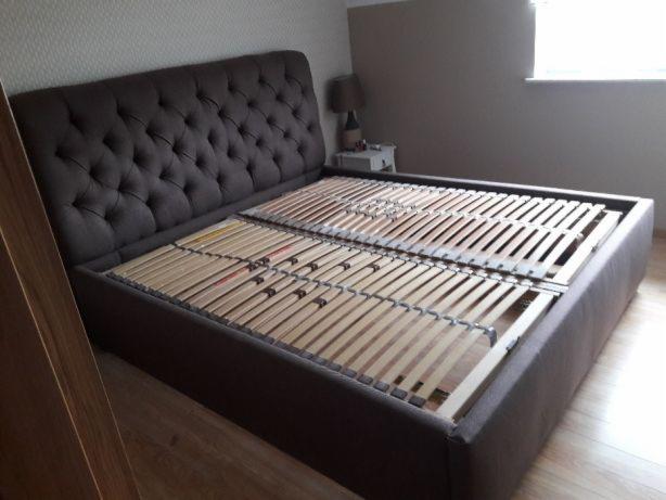 Brązowe łóżko z tapicerowanym zagłówkiem w stylu Chesterfield i drewnianym stelażem, widoczne w pokoju z oknem.