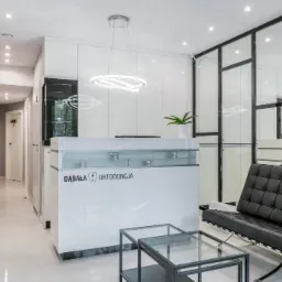 Jasne wnętrze recepcji kliniki z minimalistycznym designem, z widocznym logo firmy na ladzie recepcyjnej, nowoczesnym oświetleniem i czarną sofą w poczekalni.