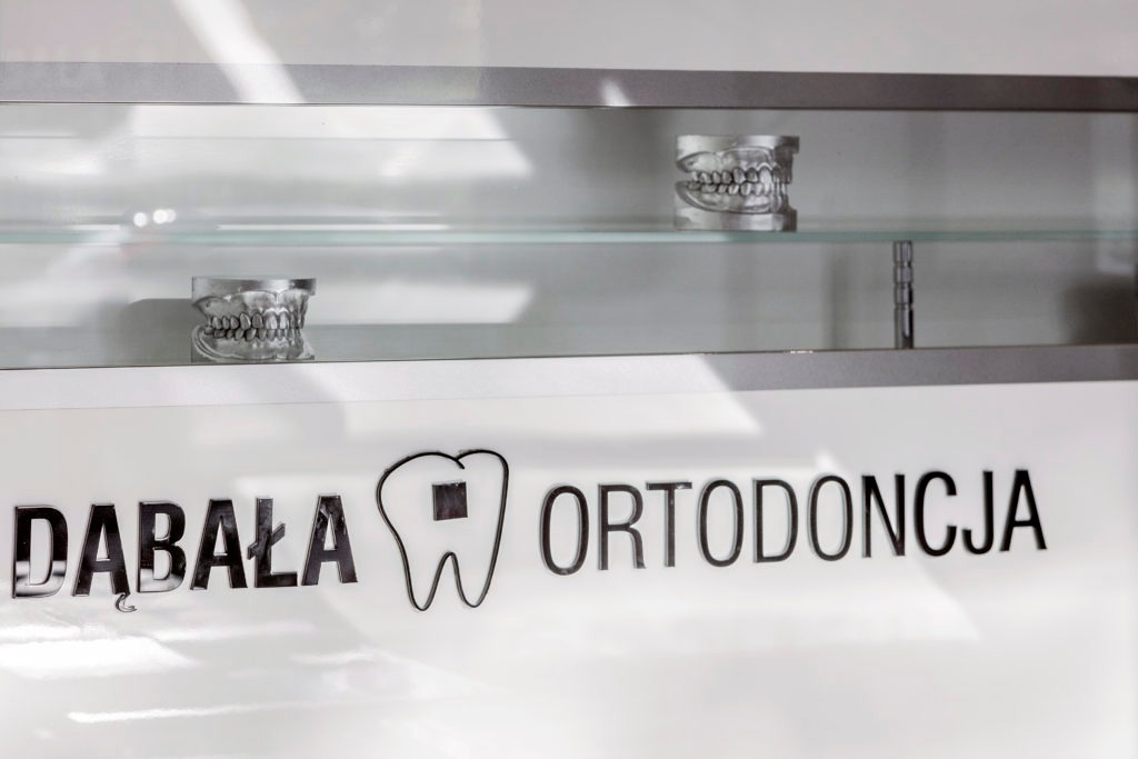 Modele szczęk z zębami na szklanej półce nad napisem 'Dąbała Ortodoncja' z logo zębów