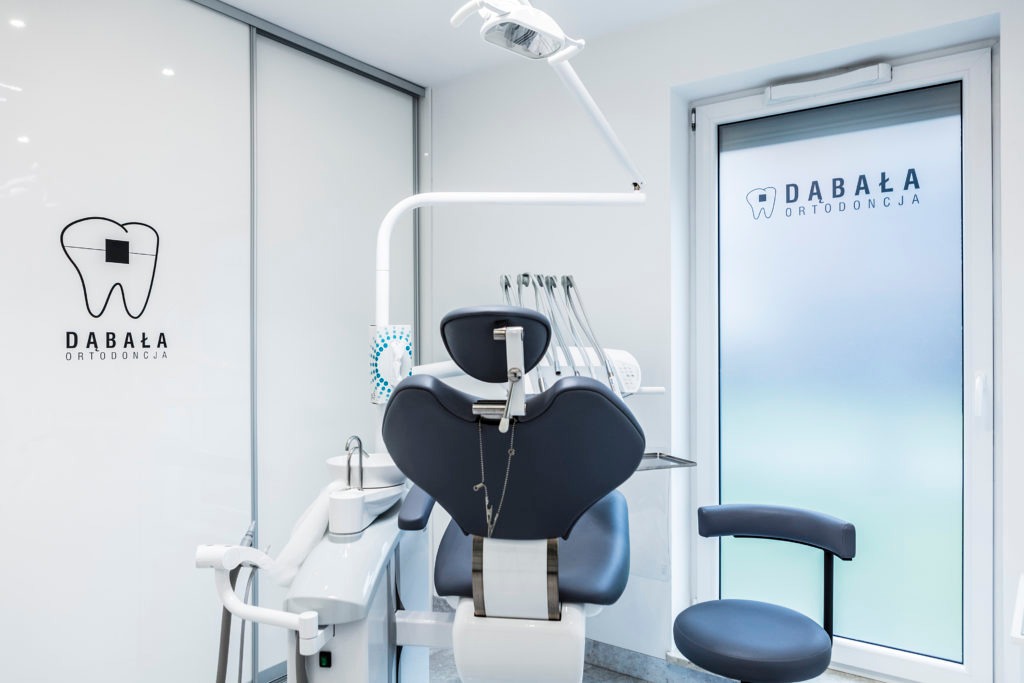 Wnętrze gabinetu lekarskiego z fotelem dentystycznym, lampą operacyjną i logo firmy na ścianie i drzwiach.