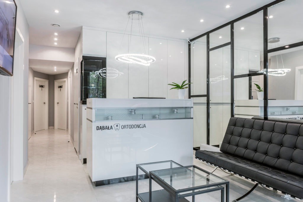 Jasne wnętrze recepcji kliniki z minimalistycznym designem, z widocznym logo firmy na ladzie recepcyjnej, nowoczesnym oświetleniem i czarną sofą w poczekalni.