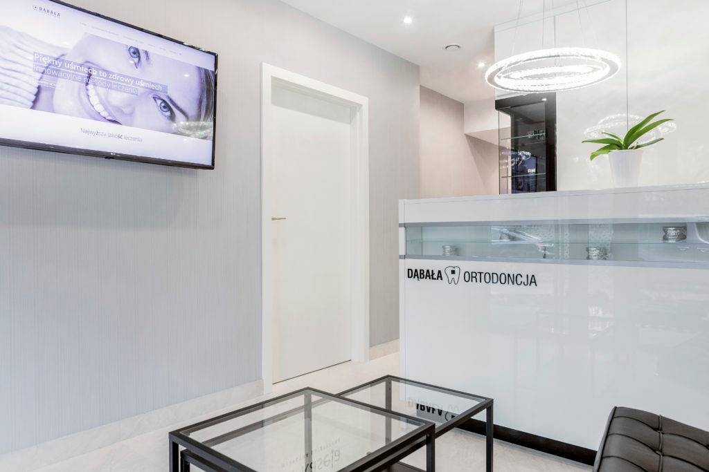 Nowoczesne wnętrze gabinetu ortodontycznego w Katowicach: biała recepcja z logo firmy, szklane stoliki kawowe, telewizor z reklamą uśmiechu i designerski żyrandol.