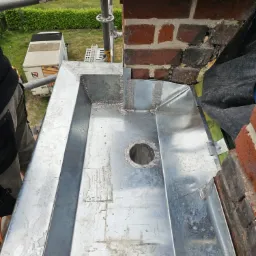 Metalowa obróbka komina z otworem, widoczna z góry podczas montażu na dachu z cegły, obok rusztowania i częściowo widocznej sylwetki pracownika.