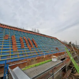 Częściowy widok dachu z czerwoną dachówką podczas montażu na niebieskiej membranie, widoczne rusztowania i przygotowane deski