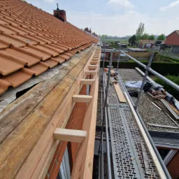 Wymiana drewnianej konstrukcji okapu dachu z czerwonej dachówki, widok z rusztowania na tle zabudowy jednorodzinnej.