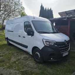 Biały samochód dostawczy Renault Master zaparkowany na trawie przed domem z panelami słonecznymi na dachu w słoneczny dzień.