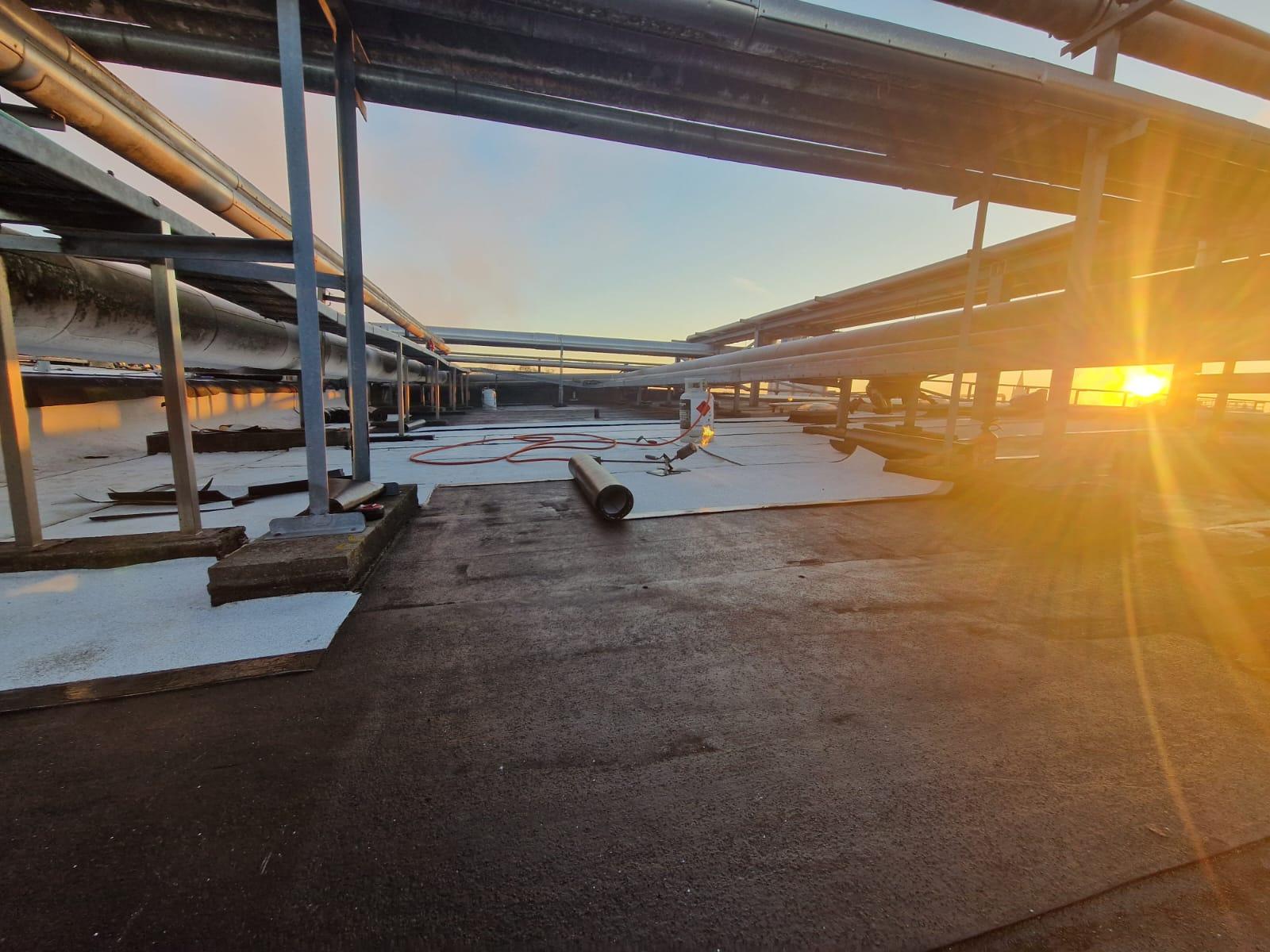 Prace na dachu płaskim: układanie nowej izolacji bitumicznej, widoczny palnik gazowy i rolka papy w promieniach zachodzącego słońca, metalowa konstrukcja nad dachem.