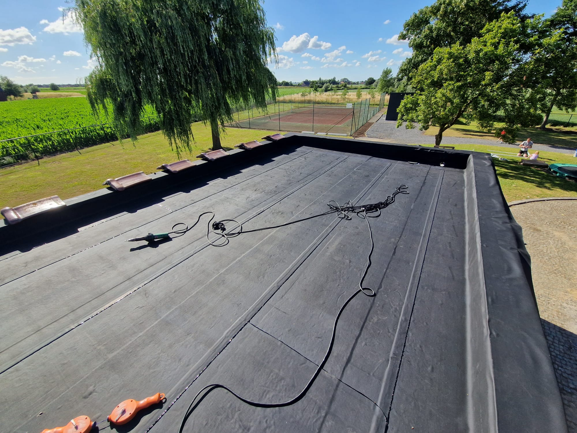 Czarny, płaski dach pokryty papą termozgrzewalną, z widocznymi narzędziami dekarskimi i fragmentem krajobrazu z drzewami i kortem tenisowym w tle.