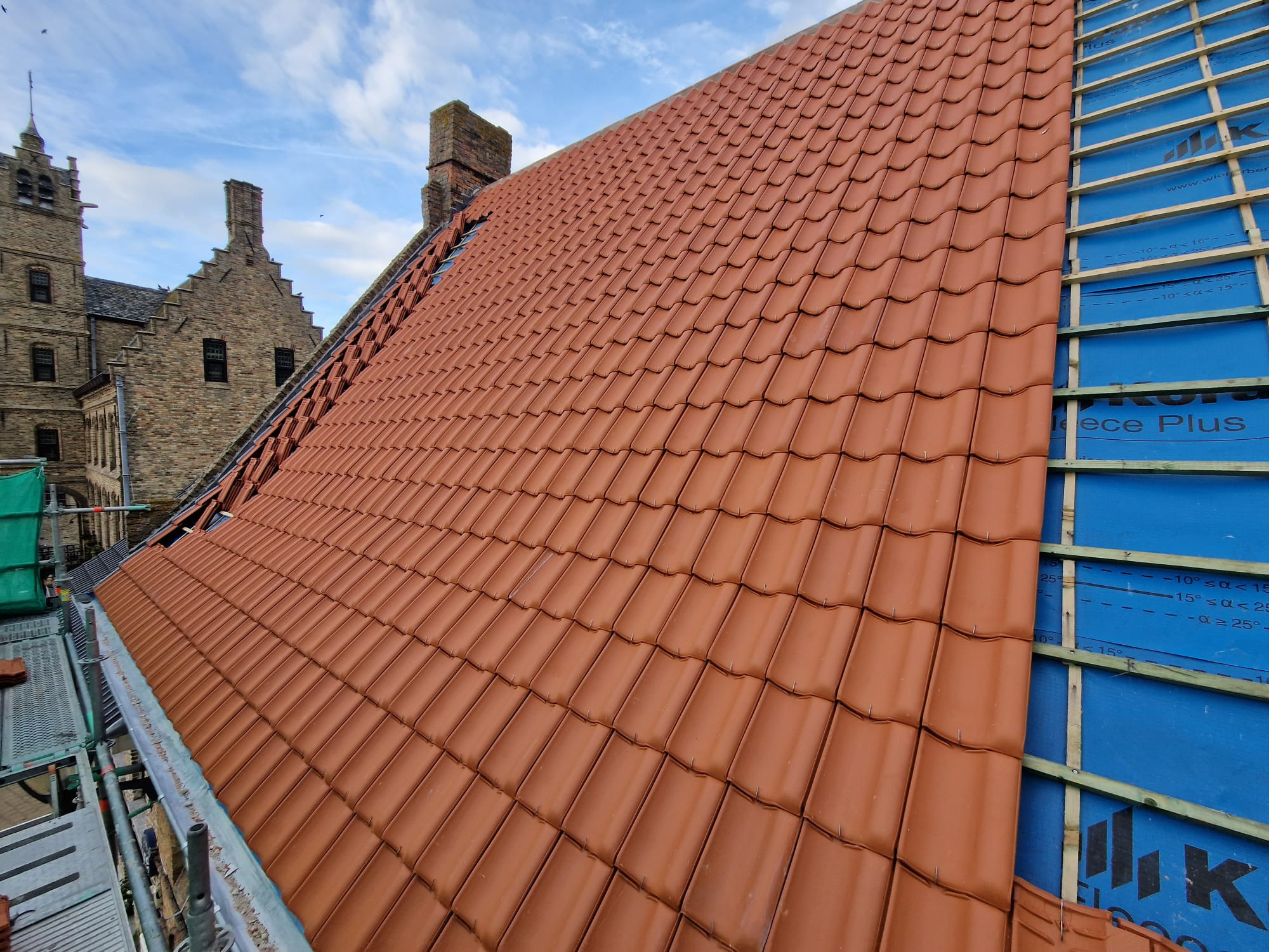 Częściowo pokryty dach z czerwonej dachówki ceramicznej, widoczna membrana izolacyjna i rusztowanie, w tle zabytkowy budynek z cegły i błękitne niebo.