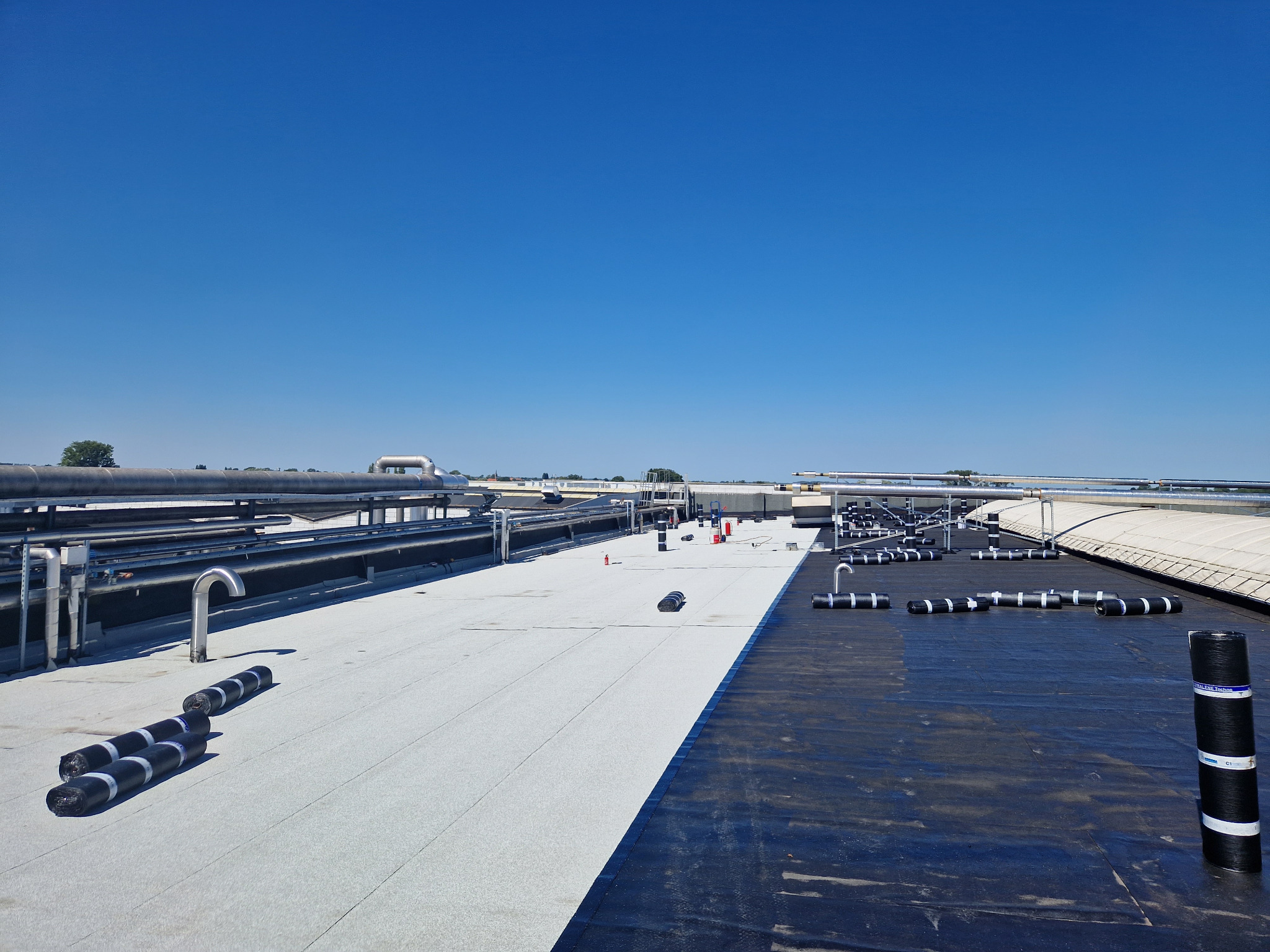 Biało-czarne krycie dachu przemysłowego z widocznymi rolkami papy na tle błękitnego nieba i systemem rur wentylacyjnych.