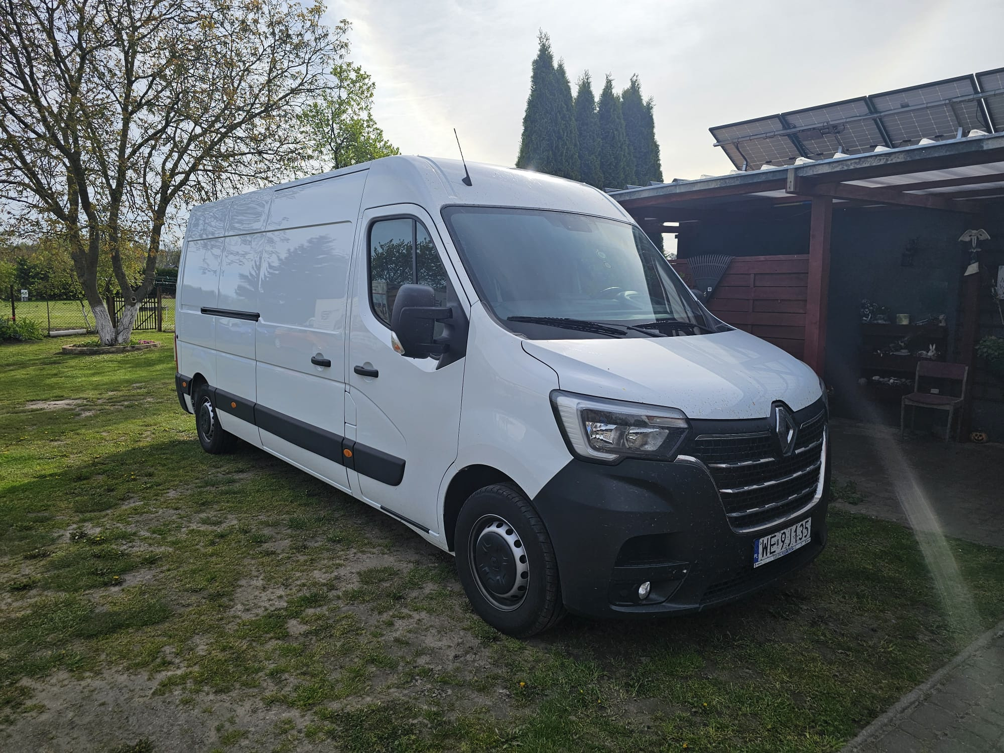 Biały samochód dostawczy Renault Master zaparkowany na trawie przed domem z panelami słonecznymi na dachu w słoneczny dzień.