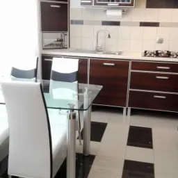 Nowoczesna kuchnia z ciemnymi drewnianymi szafkami z aluminiowymi ramkami, białymi krzesłami z czarnym akcentem przy szklanym stole z chromowanymi nogami oraz biało-czarną podłogą w szachownicę.