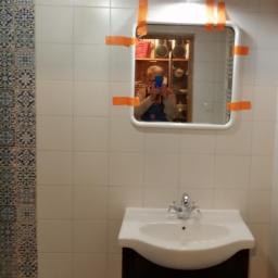 Włodzimierz Lady Remonty I Wykończenia - Łazienka w trakcie wykańczania: białe płytki, dekoracyjny pas płytek wzorzystych, umywalka z szafką, lustro z taśmą malarską, odbicie osoby robiącej zdjęcie telefonem.