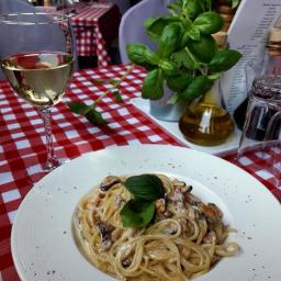 MG Dolce Vita Sp. z o.o. - Spaghetti z owocami morza na białym talerzu, kieliszek białego wina i bazylia na stole w restauracji z obrusem w czerwoną kratę.