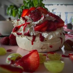 MG Dolce Vita Sp. z o.o. - Delikatna beza Pavlova z bitą śmietaną, świeżymi truskawkami, malinami, zielonymi winogronami i sosem owocowym, ustawiona na różowym talerzu z widocznym fragmentem wzorzystego obrusa w tle.