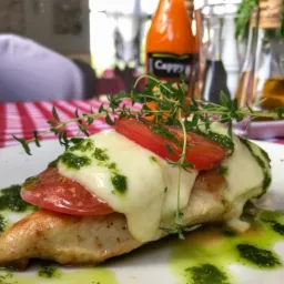 Filet z kurczaka zapiekany z serem mozzarella i pomidorem, polany pesto, udekorowany świeżym tymiankiem, na białym talerzu z czerwono-białą kratą w tle.
