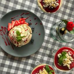 MG Dolce Vita Sp. z o.o. - Pavlova z truskawkami i borówkami na szarym talerzu, desery w szklanych pucharkach z czerwoną polewą, migdałami i miętą, oraz dekoracja z czekolady i czerwonego tulipana na tle szarej kraty.