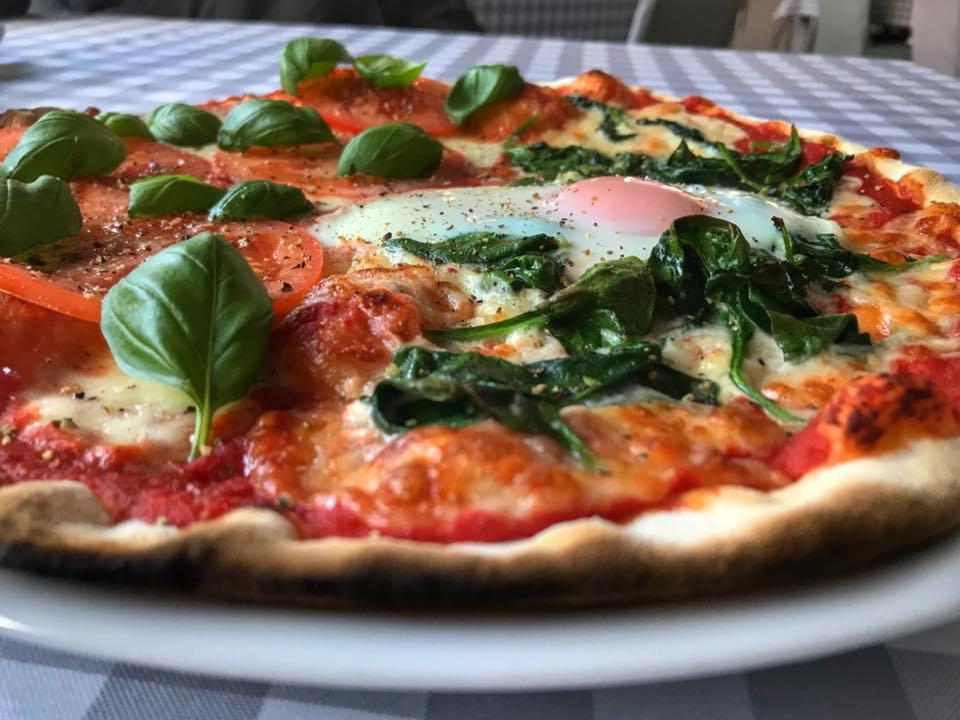 Pizza z pomidorami, szpinakiem i jajkiem sadzonym, udekorowana świeżą bazylią na talerzu z obrusem w kratę.