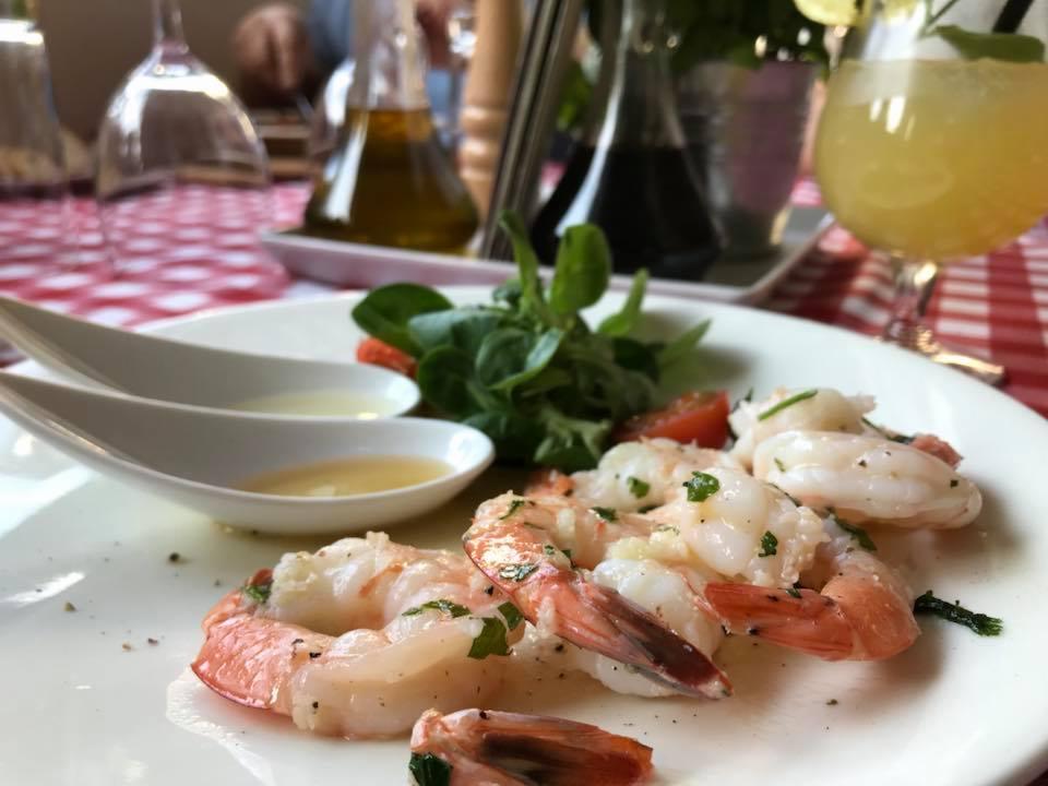 Elegancko podane krewetki z sosem i sałatką na białym talerzu, w tle napój i butelki oliwy na stole w czerwoną kratę.