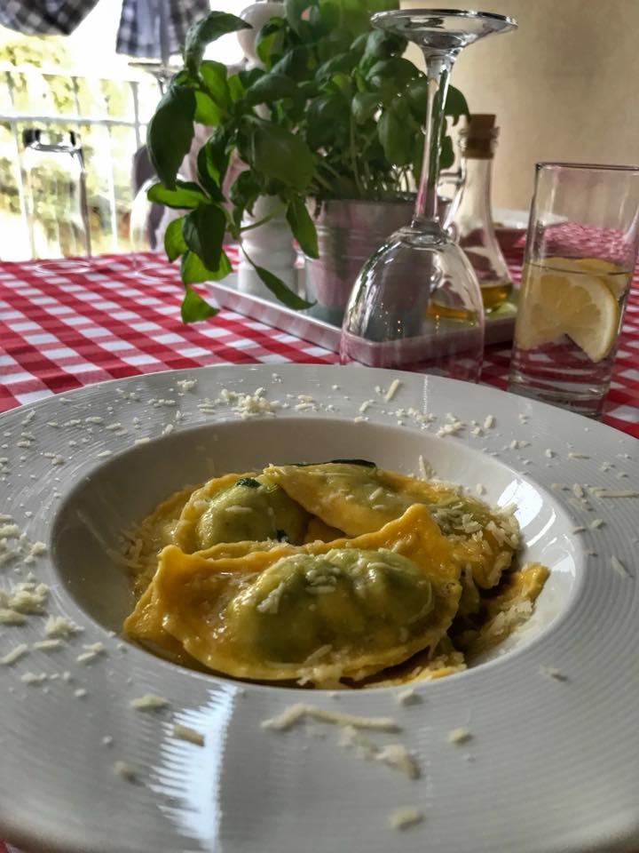 Porcja ravioli ze szpinakiem i serem, posypana parmezanem, na tle czerwono-białej kraty, z bazylią w doniczce i szklanką wody z cytryną.