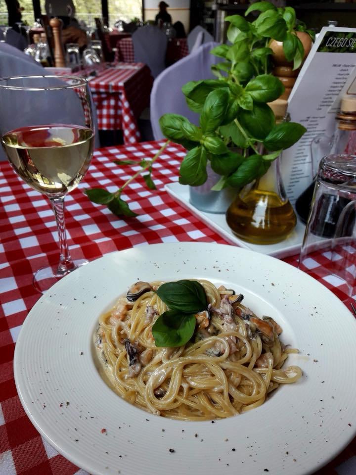 Spaghetti z owocami morza na białym talerzu, kieliszek białego wina i bazylia na stole w restauracji z obrusem w czerwoną kratę.
