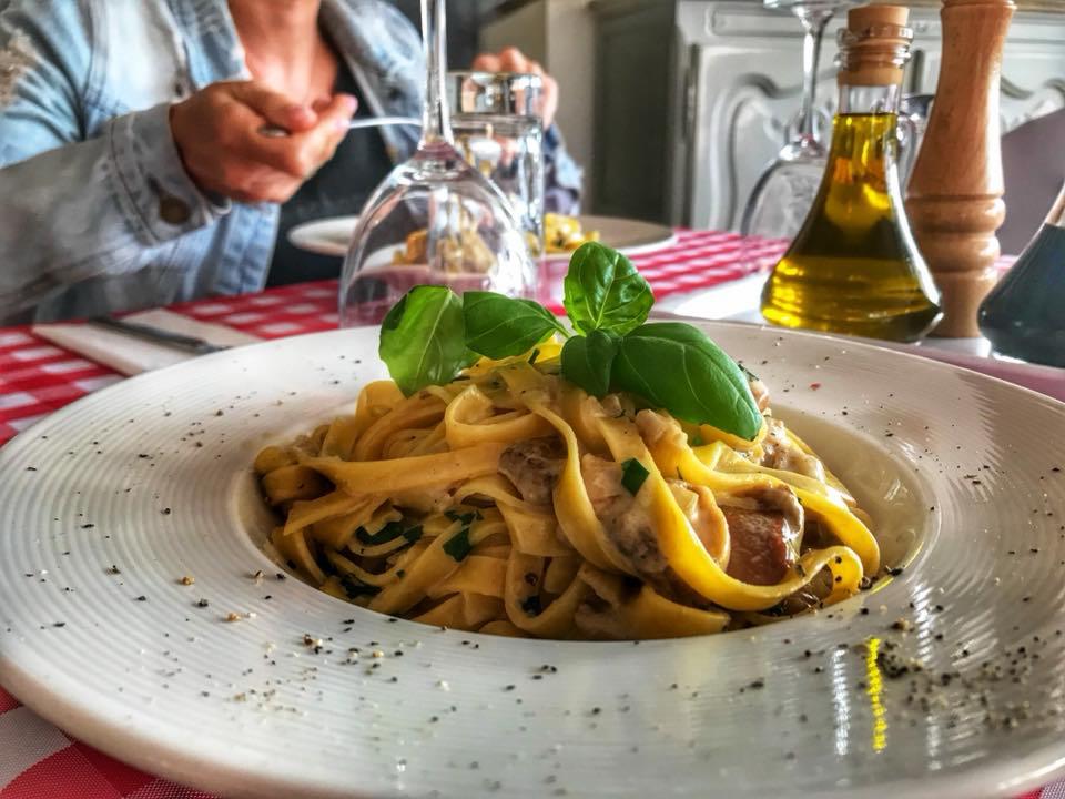 Tagliatelle z grzybami i bazylią na białym talerzu, w tle butelka oliwy i osoba jedząca, całość na czerwono-białym obrusie w kratę.