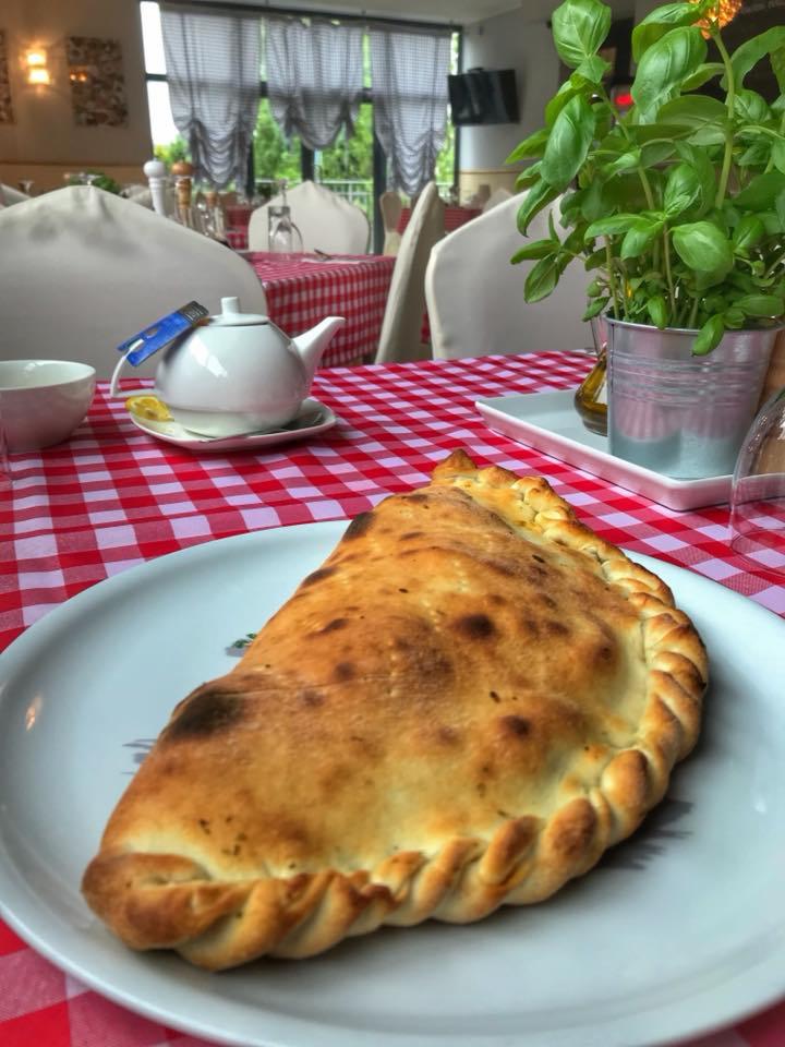 Calzone na talerzu z czerwoną kratą w tle, obok dzbanek do herbaty i doniczka z bazylią, restauracja.