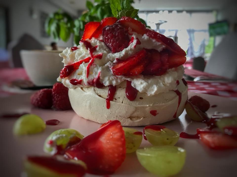 Delikatna beza Pavlova z bitą śmietaną, świeżymi truskawkami, malinami, zielonymi winogronami i sosem owocowym, ustawiona na różowym talerzu z widocznym fragmentem wzorzystego obrusa w tle.