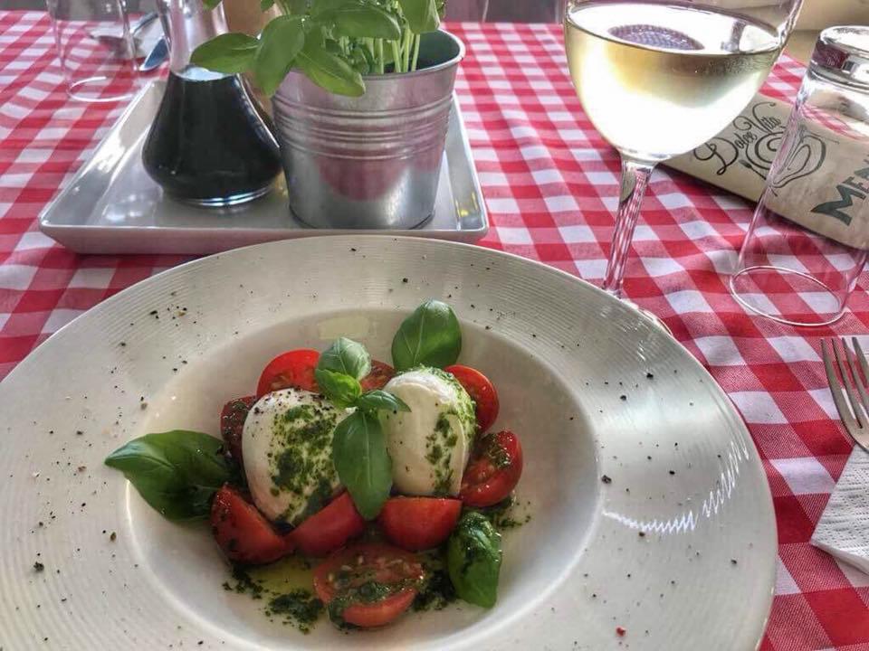 Sałatka Caprese z mozzarellą, pomidorami i bazylią na białym talerzu, podana na czerwono-białym obrusie w kratkę, obok kieliszek białego wina i doniczka z bazylią.