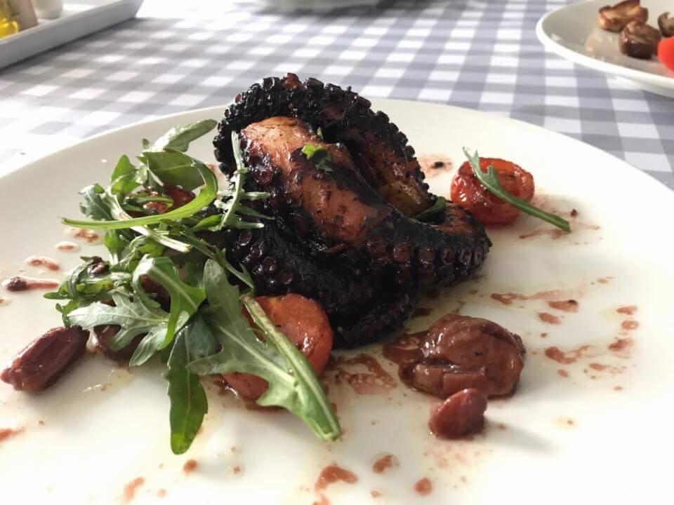 Grillowana ośmiornica podana na białym talerzu z rukolą i pomidorami cherry, na tle obrusu w kratę.