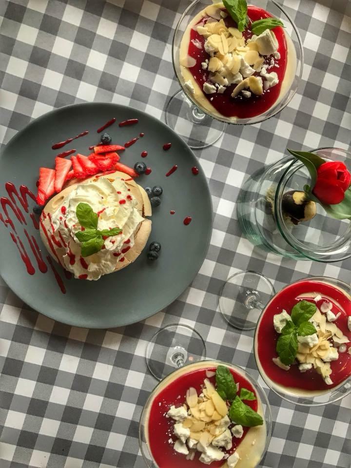 Pavlova z truskawkami i borówkami na szarym talerzu, desery w szklanych pucharkach z czerwoną polewą, migdałami i miętą, oraz dekoracja z czekolady i czerwonego tulipana na tle szarej kraty.