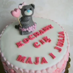 Tort urodzinowy z lukrowanym misiem trzymającym różowe serce, otoczony napisem 'Kocham Cię Maja i Julia' wykonanym z różowych literek.